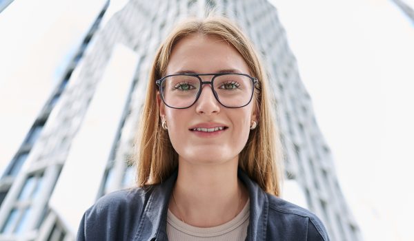 Closeup face, teenage beautiful female in glasses looking at camera, modern glass skyscrapers, urban style background. Youth, lifestyle, city life, young people concept